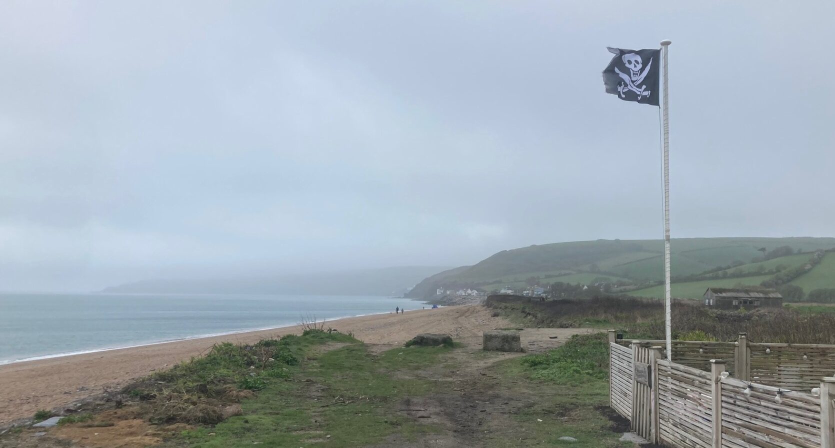 A skull and crossbones flag flying on a stretch of beach under grey skies